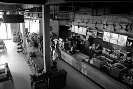 A modern, stylish coffee shop with an industrial design featuring exposed pipes and overhead lighting. The space includes a long wooden table with chairs, various coffee-making equipment, and a countertop where people are seen ordering. Shelves display various products, and light streams in from large windows, illuminating the interior. The atmosphere is lively with several patrons interacting with the baristas.