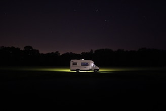 A solitary RV is parked in the middle of a dark field, illuminated by its headlights. The surrounding area is shrouded in darkness with a silhouette of trees in the background, and a clear, starry sky above.