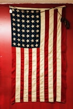 A vintage American flag displayed against a red background. The flag features alternating red and white stripes and a blue field with white stars. The fabric appears old and slightly worn, adding a sense of history.