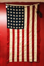 A vintage American flag displayed against a red background. The flag features alternating red and white stripes and a blue field with white stars. The fabric appears old and slightly worn, adding a sense of history.
