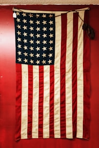 A vintage American flag displayed against a red background. The flag features alternating red and white stripes and a blue field with white stars. The fabric appears old and slightly worn, adding a sense of history.
