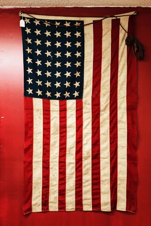A vintage American flag displayed against a red background. The flag features alternating red and white stripes and a blue field with white stars. The fabric appears old and slightly worn, adding a sense of history.
