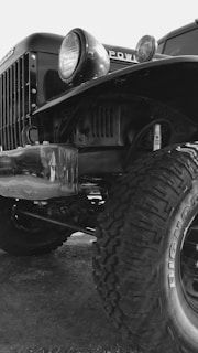 A close-up view of the front part of an old, rugged truck with large, off-road tires. The image features the truck's headlight, grille, and part of the chassis. The tire tread is prominently visible, emphasizing its heavy-duty nature.