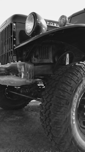 A close-up view of the front part of an old, rugged truck with large, off-road tires. The image features the truck's headlight, grille, and part of the chassis. The tire tread is prominently visible, emphasizing its heavy-duty nature.