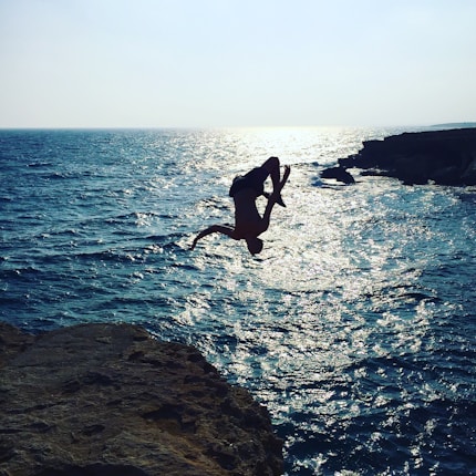 A person is performing a backflip off a cliff into the ocean. The sun glistens on the water, creating a silhouette of the person against the bright sea. Rocky cliffs surround the area, framing the scene with a sense of adventure and excitement.