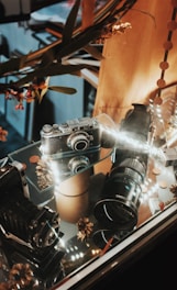 A vintage camera is displayed inside a glass case alongside a large camera lens and other photographic equipment. The setup is adorned with decorative elements, such as dried flowers and pine cones, under warm, soft lighting.