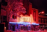 A vibrant and retro-style theater with a bright, neon-lit marquee displaying upcoming performances. Snow covers the ground and the branches of the surrounding trees, adding a wintry atmosphere. The building features a prominent sign with distinctive typography and is illuminated by red, pink, and blue lights.