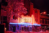 A vibrant and retro-style theater with a bright, neon-lit marquee displaying upcoming performances. Snow covers the ground and the branches of the surrounding trees, adding a wintry atmosphere. The building features a prominent sign with distinctive typography and is illuminated by red, pink, and blue lights.