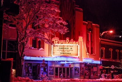 A vibrant and retro-style theater with a bright, neon-lit marquee displaying upcoming performances. Snow covers the ground and the branches of the surrounding trees, adding a wintry atmosphere. The building features a prominent sign with distinctive typography and is illuminated by red, pink, and blue lights.