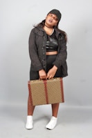 A person stands confidently, dressed in a black outfit with a jacket, cap, and sneakers. They hold a checkered suitcase with vertical brown straps. The background is neutral gray, highlighting the subject's casual and streetwear style.