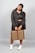 A person stands confidently, dressed in a black outfit with a jacket, cap, and sneakers. They hold a checkered suitcase with vertical brown straps. The background is neutral gray, highlighting the subject's casual and streetwear style.