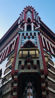 A detailed close-up of an architectural structure featuring a combination of red brick and decorative tiles with vibrant patterns. The building showcases an elaborate, geometric design with intricate textures and colors. A sky-blue background enhances the visual appeal of the unique facade.
