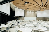 A large, modern conference room with a circular arrangement of tables and chairs. The room features a distinctive wood-paneled ceiling with embedded lights, and several large projection screens hanging from the ceiling. A camera and equipment are set up in the middle of the room.