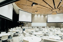 A large, modern conference room with a circular arrangement of tables and chairs. The room features a distinctive wood-paneled ceiling with embedded lights, and several large projection screens hanging from the ceiling. A camera and equipment are set up in the middle of the room.