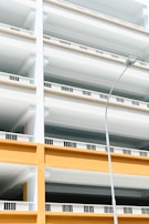 A modern, multi-level parking structure with clean, geometric lines and open railings. The facade features alternating sections of white and bright yellow-orange, with a single streetlight in the foreground.