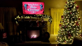 A cozy living room decorated for Christmas, featuring a large, illuminated Christmas tree adorned with ornaments and lights. A fireplace is surrounded by a garland with stockings hanging above. A television above the fireplace displays a festive movie title. Soft lighting and decorative elements enhance the warm holiday atmosphere.