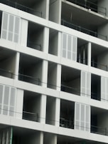 A partially constructed building featuring multiple rectangular openings and unfinished concrete walls. Metal railings are installed across each level, and some window frames have been fitted with ventilation or decorative panels.