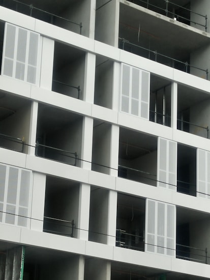 A partially constructed building featuring multiple rectangular openings and unfinished concrete walls. Metal railings are installed across each level, and some window frames have been fitted with ventilation or decorative panels.
