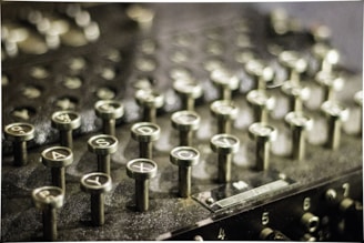 A close-up image of an old mechanical machine with multiple circular keys featuring letters and numbers. The surface appears to be made of metal with a vintage or antique appearance. The keys are protruding and arranged in neat rows, suggesting it might be a typewriter or an encryption device.