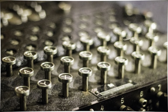 A close-up image of an old mechanical machine with multiple circular keys featuring letters and numbers. The surface appears to be made of metal with a vintage or antique appearance. The keys are protruding and arranged in neat rows, suggesting it might be a typewriter or an encryption device.