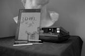 A black and white photograph featuring a Polaroid camera placed on a dark fabric surface, accompanied by two developing photographs and a book titled 'IDOL' with an image of a classical sculpture on its cover. In the background, there is a bust of a statue.