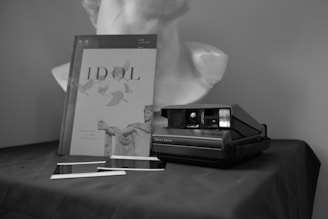 A black and white photograph featuring a Polaroid camera placed on a dark fabric surface, accompanied by two developing photographs and a book titled 'IDOL' with an image of a classical sculpture on its cover. In the background, there is a bust of a statue.