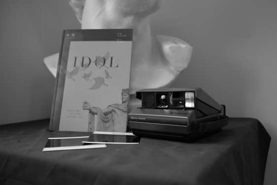 A black and white photograph featuring a Polaroid camera placed on a dark fabric surface, accompanied by two developing photographs and a book titled 'IDOL' with an image of a classical sculpture on its cover. In the background, there is a bust of a statue.