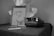 A black and white photograph featuring a Polaroid camera placed on a dark fabric surface, accompanied by two developing photographs and a book titled 'IDOL' with an image of a classical sculpture on its cover. In the background, there is a bust of a statue.