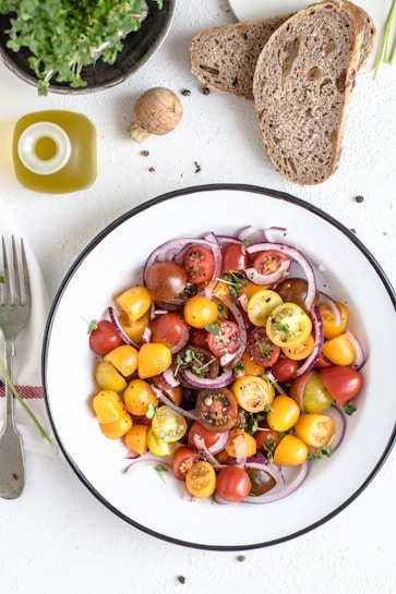 A fresh salad featuring a variety of colorful cherry tomatoes, sliced red onions, and green herbs. The salad is served in a white bowl with a black rim. Accompanying the salad are slices of whole-grain bread, a small green bottle of olive oil, and a wooden cork. A black potted green plant is placed nearby, adding a touch of nature to the scene. A fork and a striped napkin are also present beside the plate.