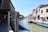 A canal lined with colorful buildings is bustling with activity, featuring small boats docked along the sides. People are walking along the waterfront, and the architecture showcases a blend of historic and vibrant facades. In the distance, a small bridge arches over the canal.