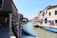 A canal lined with colorful buildings is bustling with activity, featuring small boats docked along the sides. People are walking along the waterfront, and the architecture showcases a blend of historic and vibrant facades. In the distance, a small bridge arches over the canal.