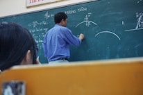 A teacher is standing at a chalkboard, writing equations and diagrams. The board contains various mathematical notations and illustrations related to physics. A student appears to be observing attentively while taking notes.