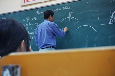 A teacher is standing at a chalkboard, writing equations and diagrams. The board contains various mathematical notations and illustrations related to physics. A student appears to be observing attentively while taking notes.