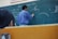 A teacher is standing at a chalkboard, writing equations and diagrams. The board contains various mathematical notations and illustrations related to physics. A student appears to be observing attentively while taking notes.