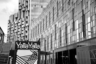 High-rise buildings with modern architectural designs dominate the scene. The structures feature numerous rectangular glass panels, creating a reflective façade. At the foreground, a sign with the text 'Yalla Yalla' is prominently displayed, accompanied by abstract black lines on a white background.