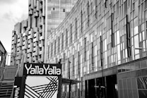 High-rise buildings with modern architectural designs dominate the scene. The structures feature numerous rectangular glass panels, creating a reflective fa&ccedil;ade. At the foreground, a sign with the text 'Yalla Yalla' is prominently displayed, accompanied by abstract black lines on a white background.
