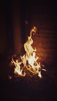 Bright, dancing flames flicker upwards from a pile of what appears to be charred material, illuminating the surrounding darkness with a warm, yellow-orange glow. The rusty surface in the background suggests an industrial setting.