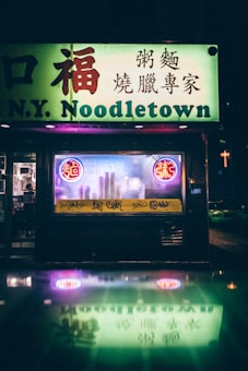 A neon-lit storefront of a noodle shop with signage in both English and Chinese characters. Bright neon colors illuminate the dark street, creating a reflected glow on a wet surface. Two circular neon signs add a vibrant touch, while the shop's window shows urban graffiti.