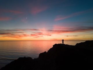 A joyful traveler standing on a scenic cliff overlooking a vibrant sunset over the ocean.
