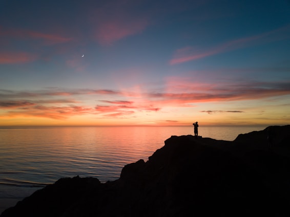 A joyful traveler standing on a scenic cliff overlooking a vibrant sunset over the ocean.