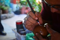 A person is intricately carving or inscribing on a round brown object, possibly a gourd or wooden item, using a sharp tool. Their hands are focused, and the environment suggests a crafting or artistic workshop with blurred plants and tools in the background.
