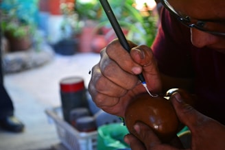 A person is intricately carving or inscribing on a round brown object, possibly a gourd or wooden item, using a sharp tool. Their hands are focused, and the environment suggests a crafting or artistic workshop with blurred plants and tools in the background.