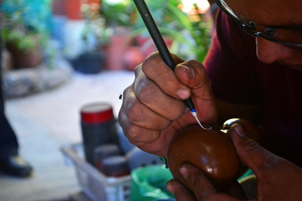 A person is intricately carving or inscribing on a round brown object, possibly a gourd or wooden item, using a sharp tool. Their hands are focused, and the environment suggests a crafting or artistic workshop with blurred plants and tools in the background.