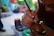 A person is intricately carving or inscribing on a round brown object, possibly a gourd or wooden item, using a sharp tool. Their hands are focused, and the environment suggests a crafting or artistic workshop with blurred plants and tools in the background.