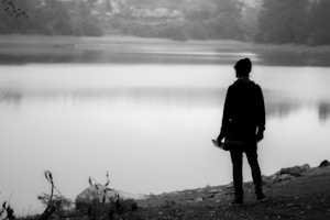 A silhouette of a person is standing near the edge of a calm body of water, gazing across it. The surrounding environment is serene and misty, with trees visible in the background. The image is in black and white, enhancing the tranquil and introspective mood. Some foliage and rocks are present on the ground.