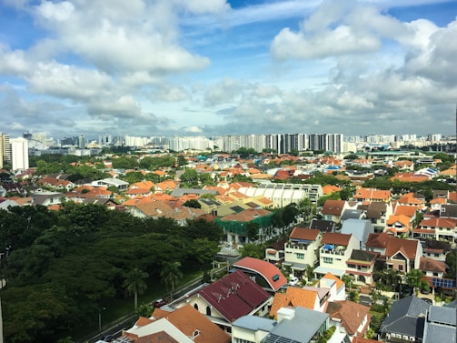 An urban landscape with a mix of residential houses and high-rise buildings. The foreground features numerous houses with red and brown tiled roofs, surrounded by lush greenery. The background includes a skyline of modern high-rise buildings and scattered clouds in a bright blue sky.