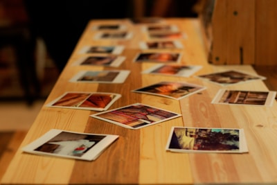 Several printed photographs are scattered across a wooden table. The photographs vary in orientation and depict different scenes or subjects, creating a nostalgic and artistic arrangement. The warm tones of the wood complement the varied colors in the photographs.