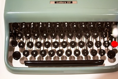A vintage typewriter keyboard with round black keys featuring white letters and symbols. The keys are arranged in a classic QWERTY layout, set against a light blue-green body with a smooth finish. The typewriter model 'Lettera 22' is indicated above the keys. A single bright red key adds a pop of color amidst the otherwise monochromatic palette.