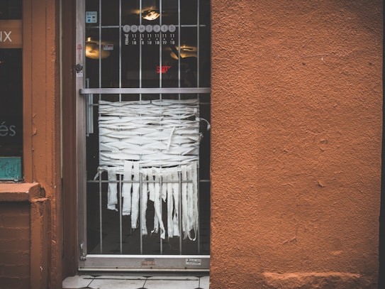 A storefront entrance with a metal grated door, behind which strips of white fabric are intricately woven. The wall next to the door is textured and painted in a rusty orange color. A partial window display is visible on the left side, with some stickers and decals.