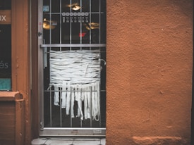 A storefront entrance with a metal grated door, behind which strips of white fabric are intricately woven. The wall next to the door is textured and painted in a rusty orange color. A partial window display is visible on the left side, with some stickers and decals.